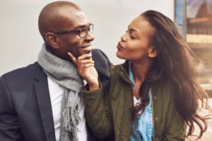 An image of a young African American woman flirtatiously pursing her lips for a kiss while gently caressing the face of a handsome man wearing glasses. They appear to be enjoying a date together. This image could be associated with signs a married woman wants to sleep with you